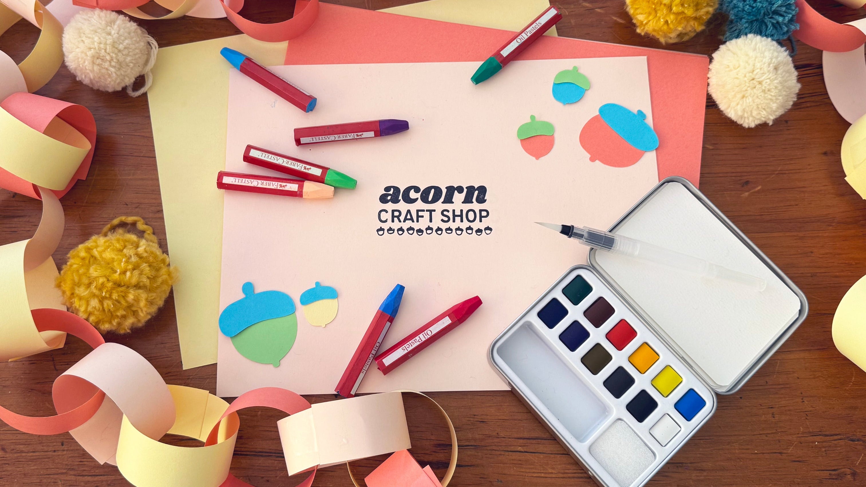 Colorful paper chain, markers, pom-poms, and a paint palette on a wooden surface with 'Acorn Craft Shop' branding.